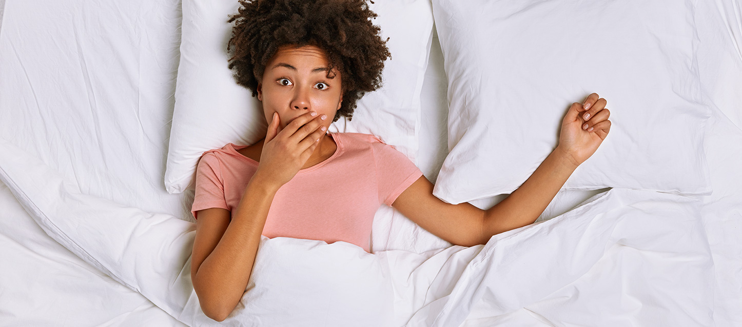 Fille brune aux longs cheveux afro noirs vêtue d’une chemise rose, couchée sur le dos dans un lit, couverte d’un édredon avec la main se couvrant sa bouche dans un geste de surprise.
