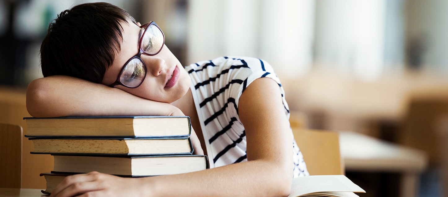 Fille aux cheveux bruns courts avec des lunettes et un T-shirt blanc avec des rayures noires, dort sur le siège d’une bibliothèque appuyé sur plusieurs livres.